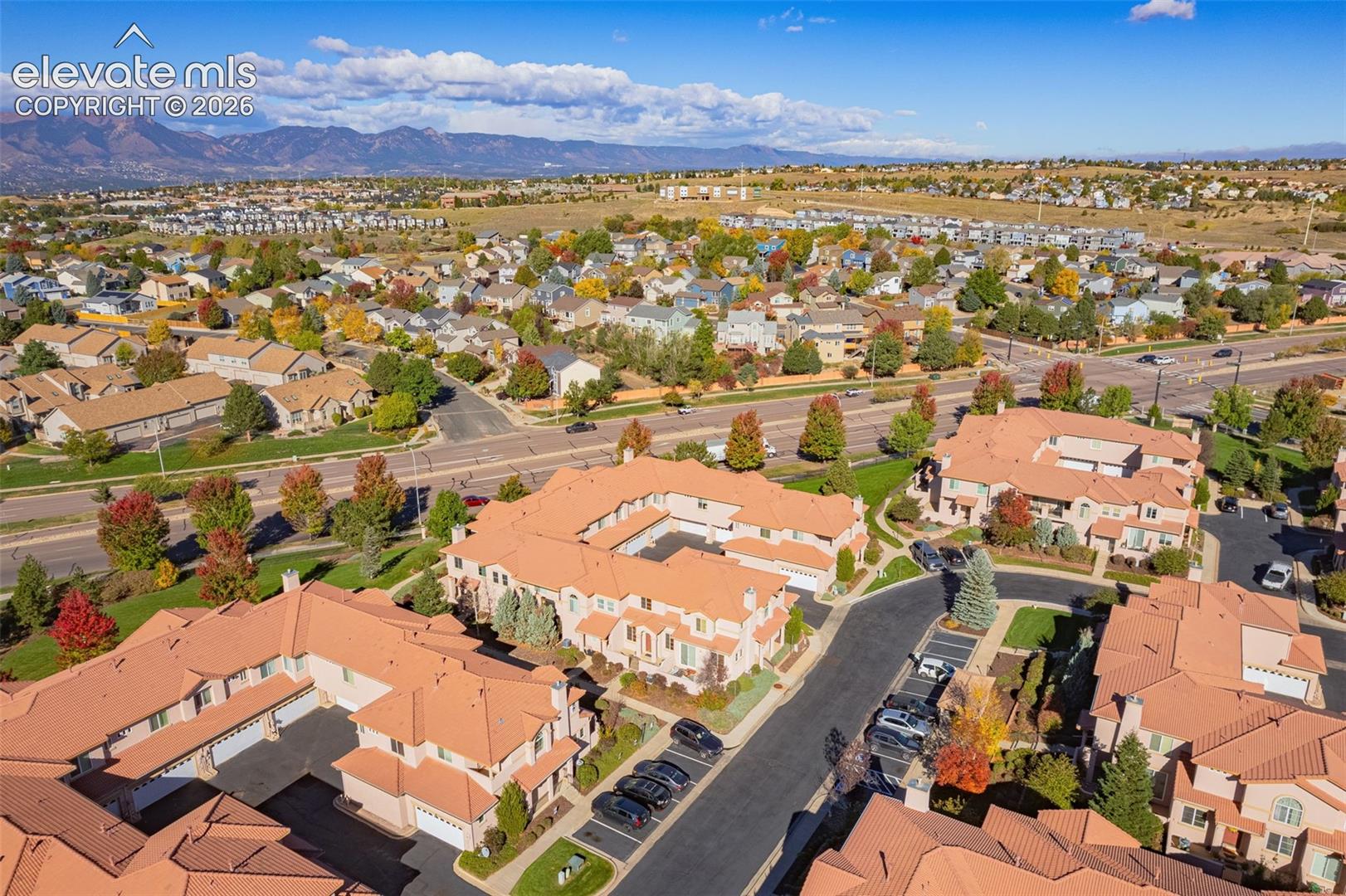 Aerial perspective of a multi-unit residential complex featuring terracotta-tiled roofs, stucco siding, paved driveway access, attached garage bays, and a mountain backdrop