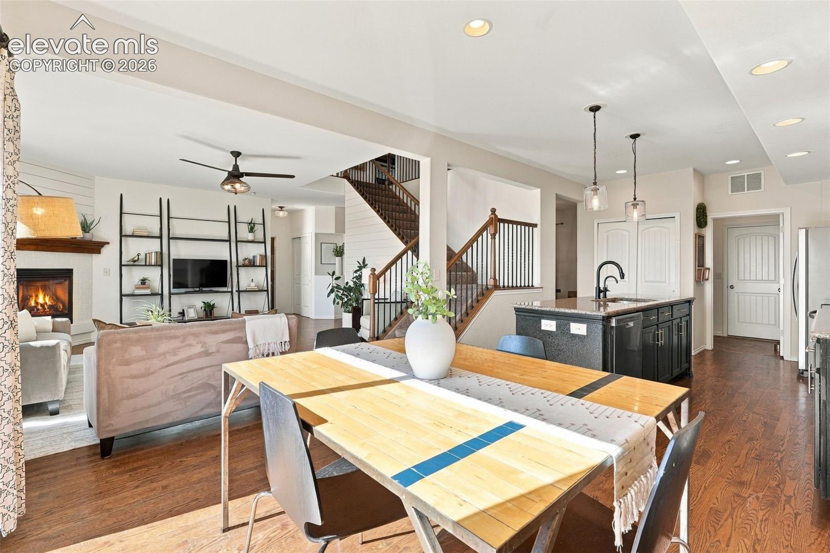 Open-concept interior featuring wood-finish flooring, a fireplace with a wooden mantel, a staircase with a stained wood handrail and vertical balusters, and a kitchen island with dark cabinetry and a stone-surface countertop