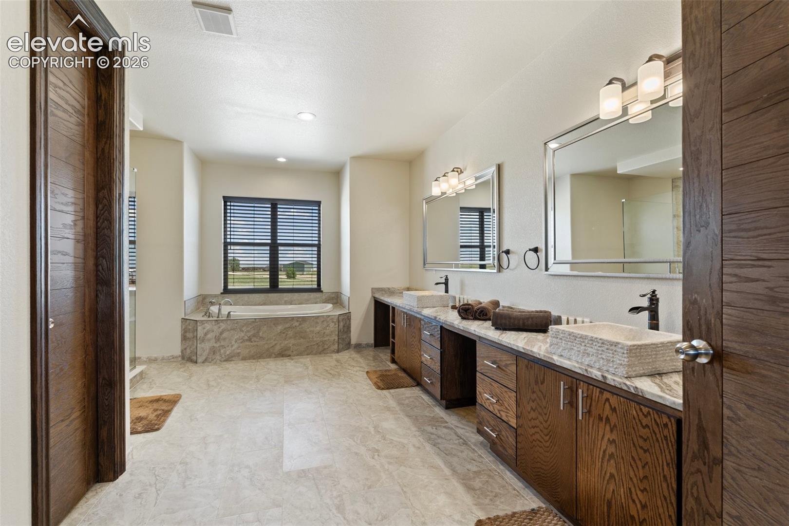 Spacious primary bath featuring a built-in soaking tub, dual vanity with vessel sinks, wood-finish cabinetry, and large format floor tile