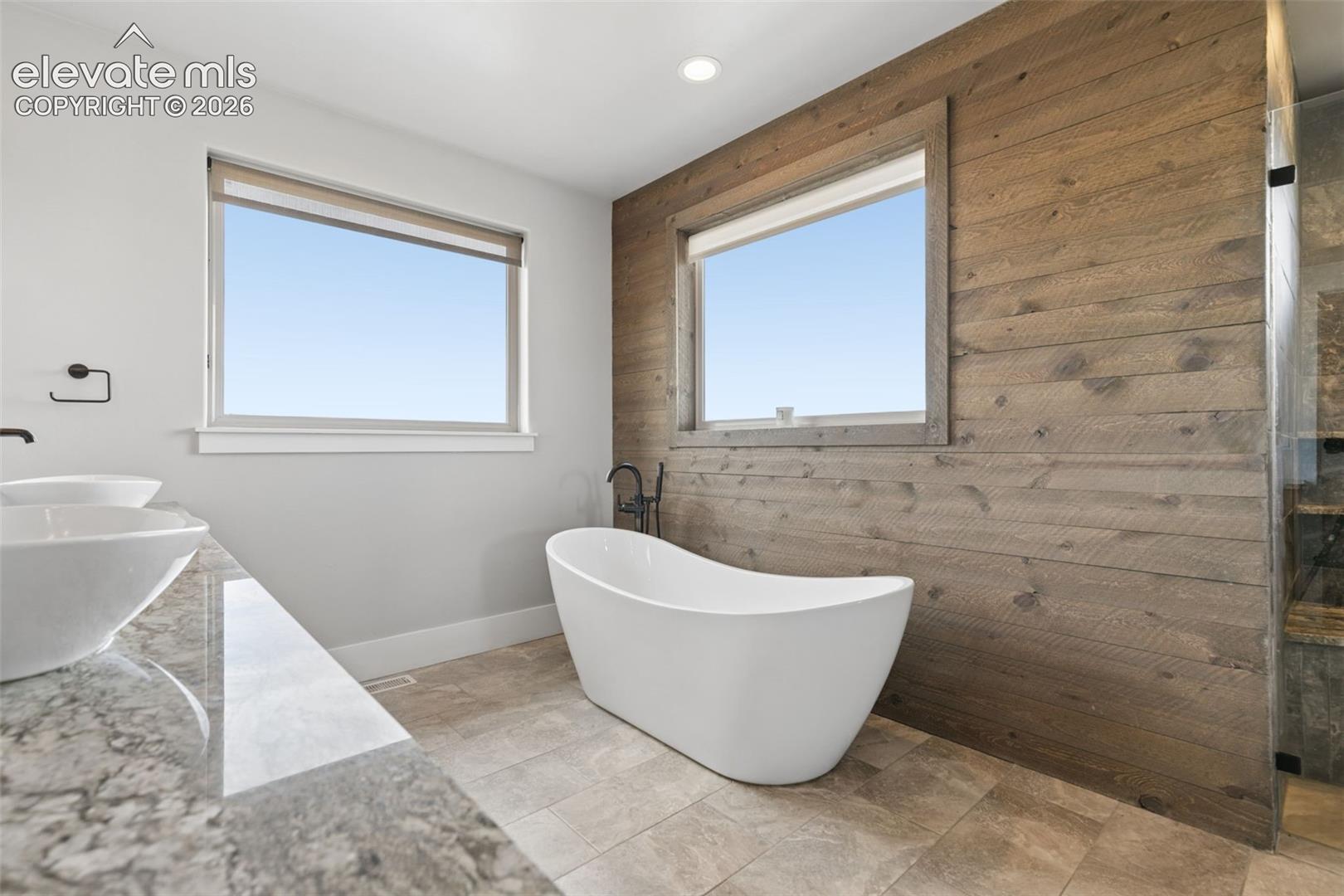 Freestanding soaking tub, wood-finish accent wall, dual vessel sinks, stone-finish vanity, and a glass-enclosed shower