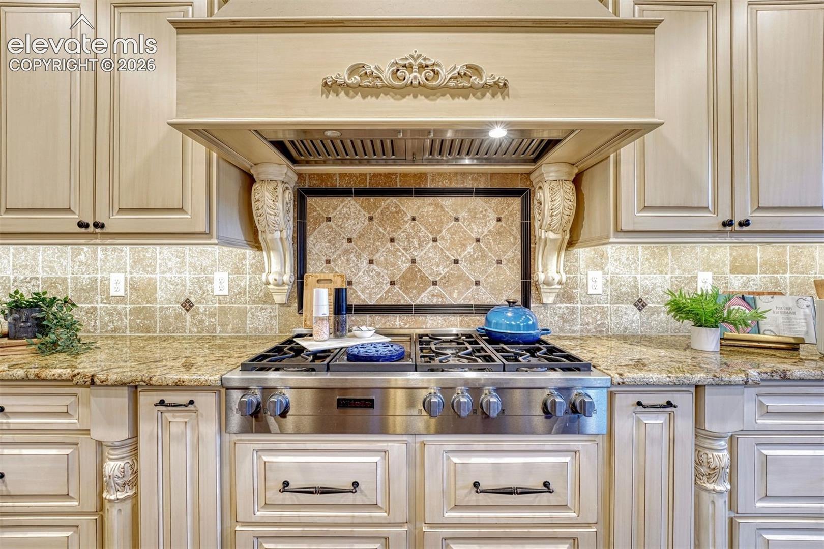 Built-in stainless steel range, custom carved wood range hood, decorative tile backsplash, light-toned cabinetry with dark hardware, and granite countertops
