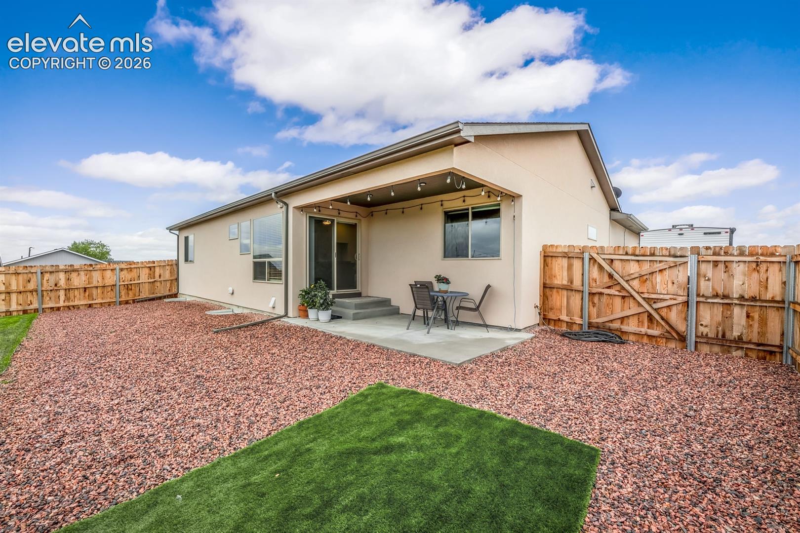 Rear exterior featuring a covered concrete patio, synthetic turf area, decorative stone ground cover, and full perimeter wood fencing