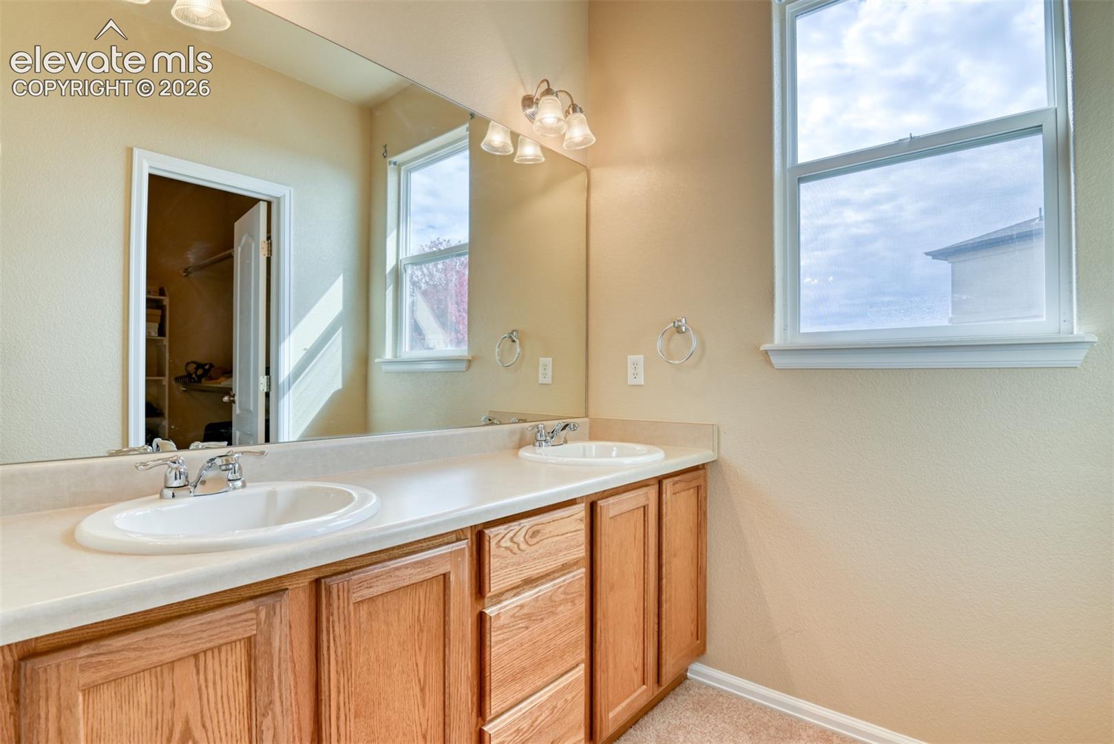 Dual-sink vanity featuring wood-finish cabinetry, solid-surface countertop, and wall-to-wall vanity mirror with multi-bulb overhead lighting