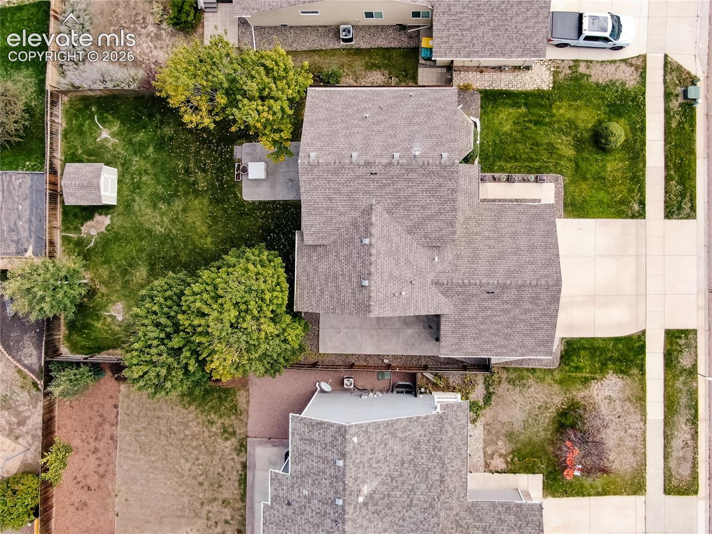 Aerial perspective of property featuring shingle roof, concrete driveway, fenced yard, detached storage structure, and mature trees