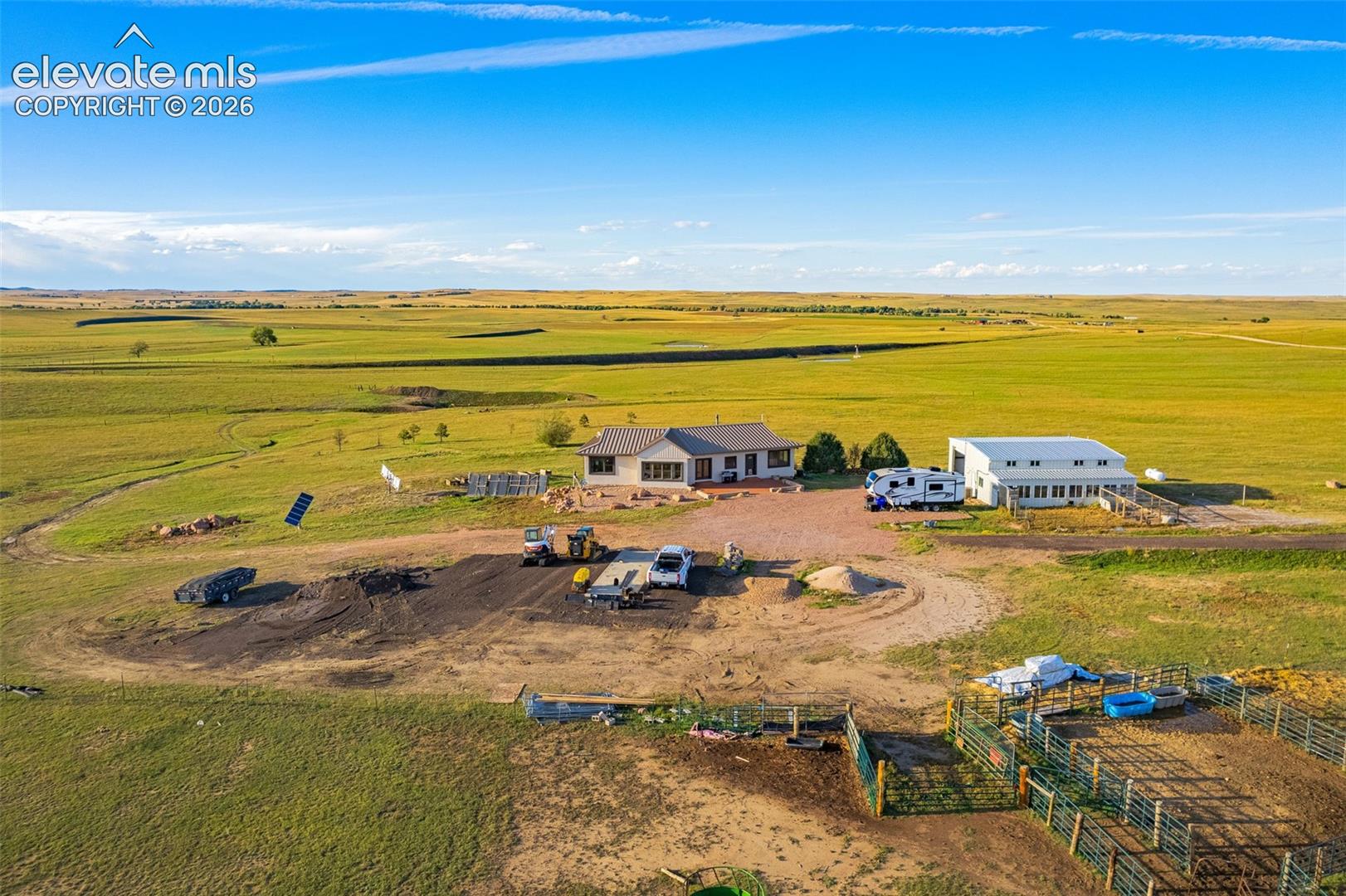 Expansive rural property featuring a primary residence with a gray roof, a large secondary building, and fenced corrals