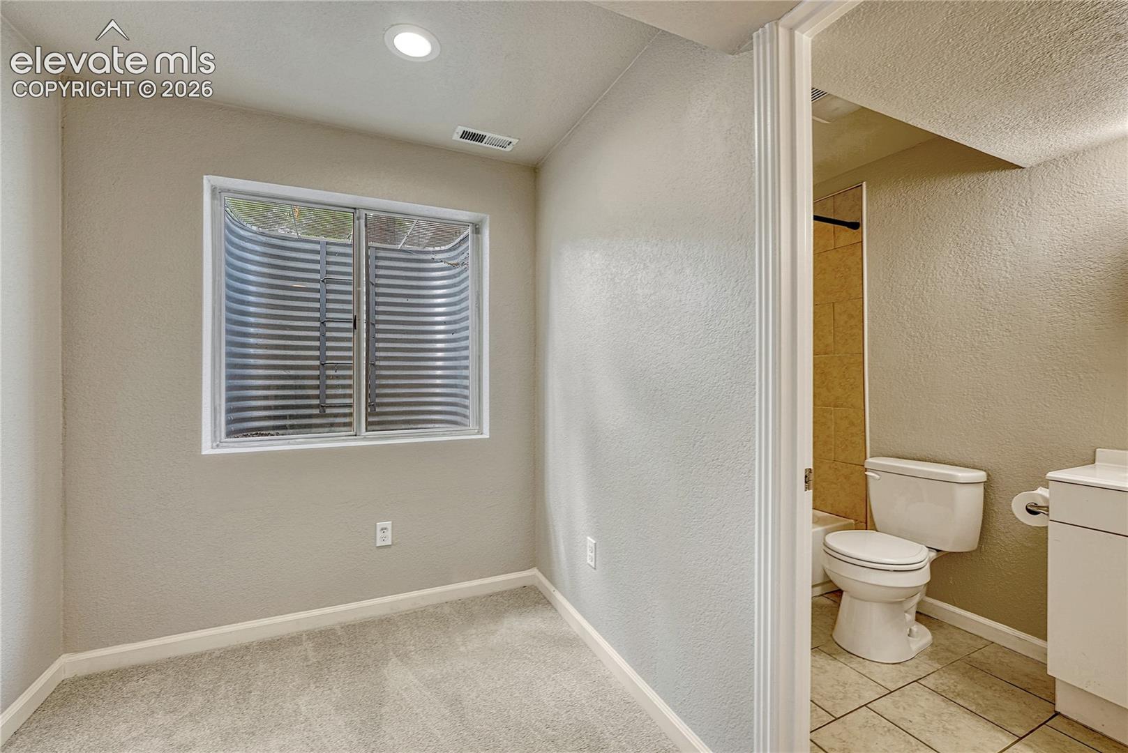 Neutral-toned room featuring wall-to-wall carpeting and a recessed window well, paired with a bathroom containing a tiled shower surround, porcelain toilet, and white vanity