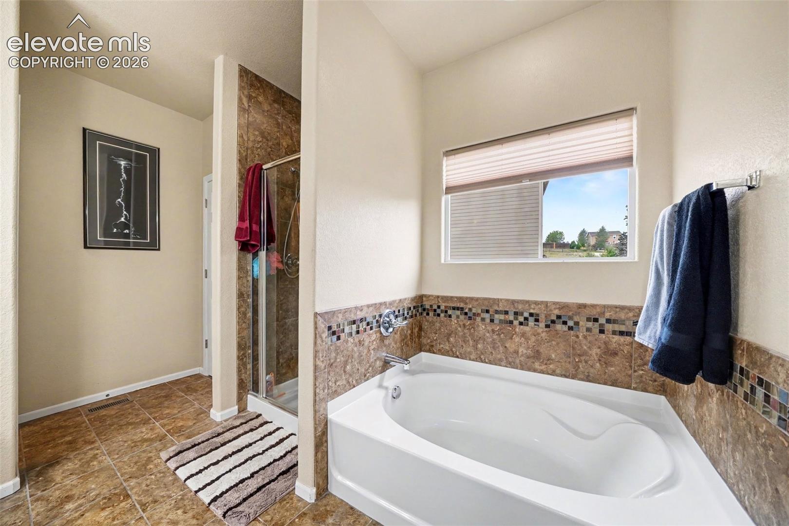 Corner soaking tub, tiled shower enclosure, large window, and tiled flooring