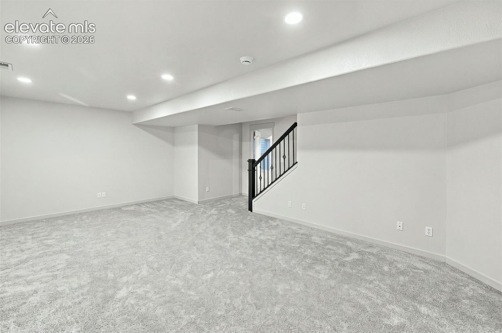 Open-concept room featuring neutral wall-to-wall carpeting, recessed ceiling lighting, and a staircase with a black metal spindle railing