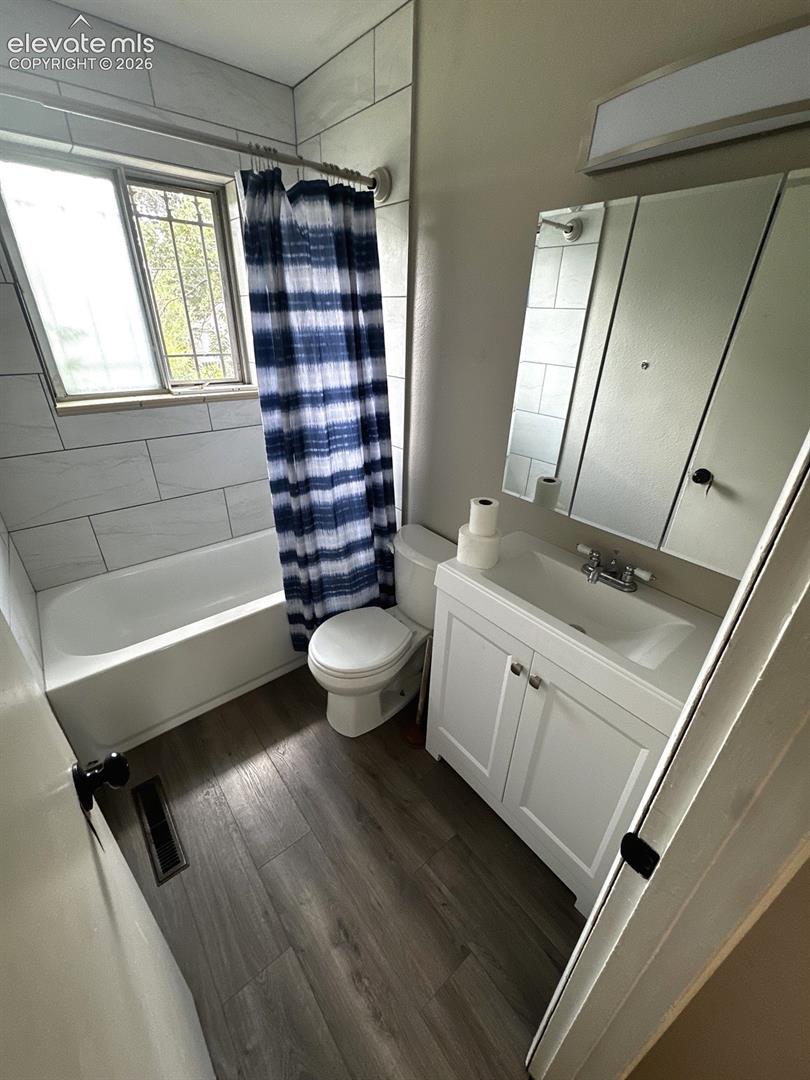 White vanity with a rectangular sink, wood-finish flooring, white subway tile shower surround, and a window with security bars
