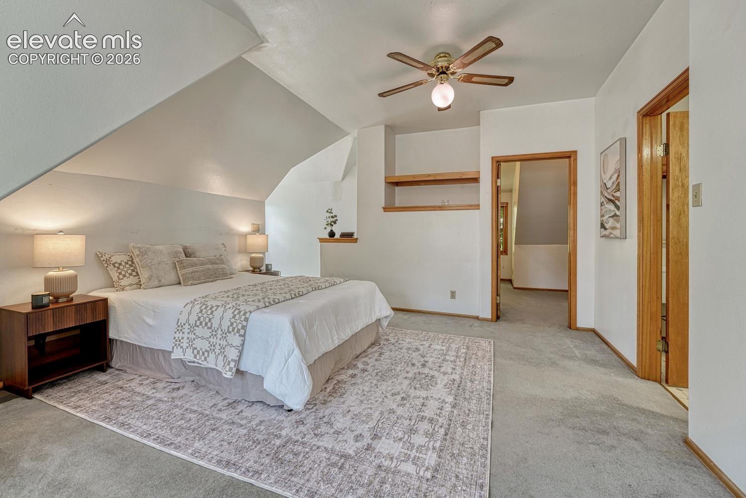 Vaulted ceiling, ceiling fan with light fixture, light wood trim, built-in shelving, and light gray carpeting
