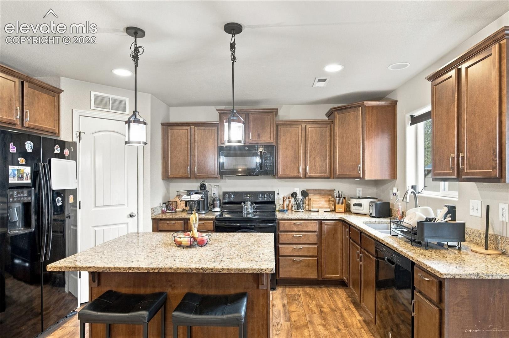 Kitchen featuring stained wood cabinetry, stone countertops, a center island with pendant lighting, and wood-finish flooring