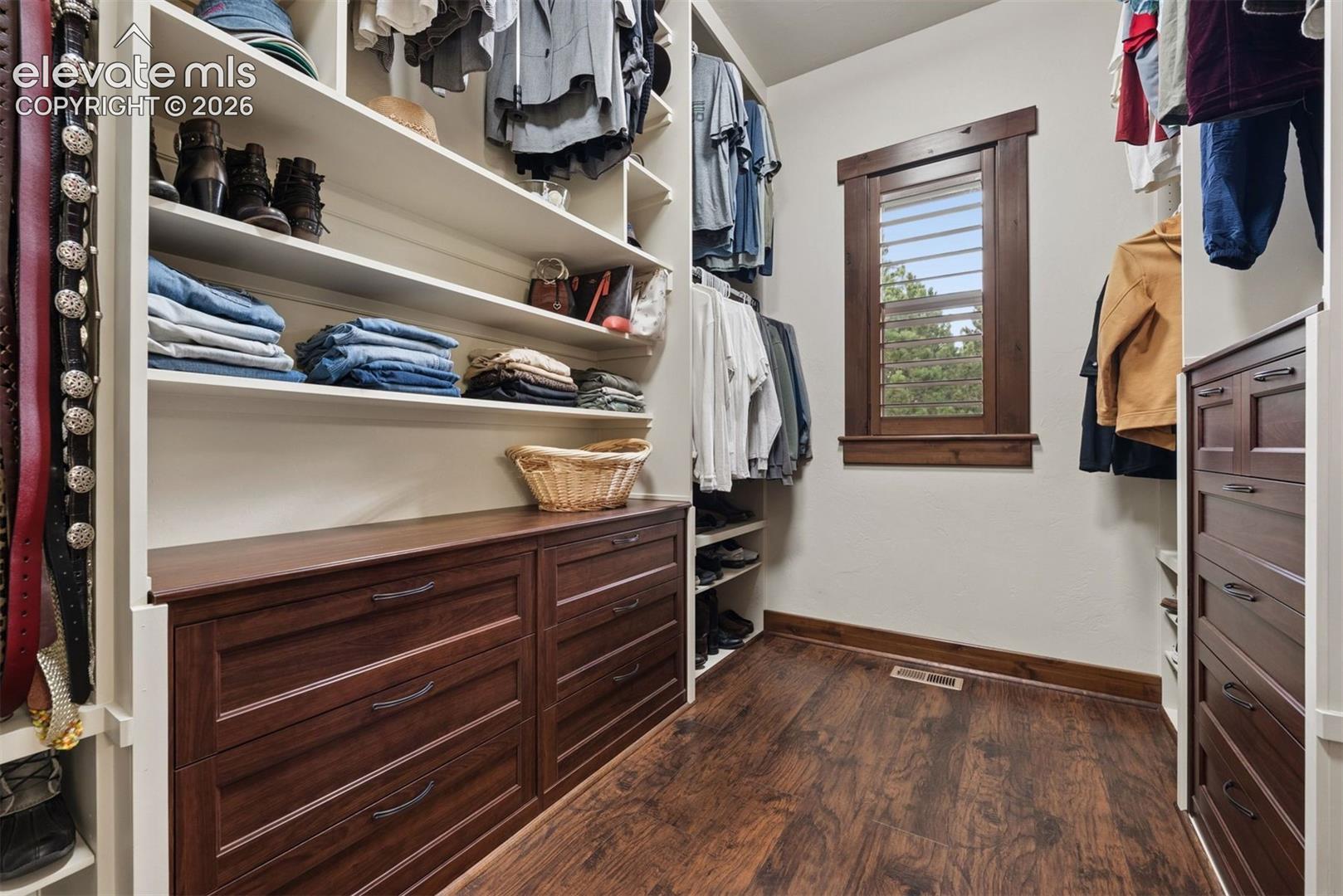 Walk-in closet featuring floor-to-ceiling built-in cabinetry with wood-finish drawers, adjustable open shelving, wood-finish flooring, and a window framed by dark-stained wood trim