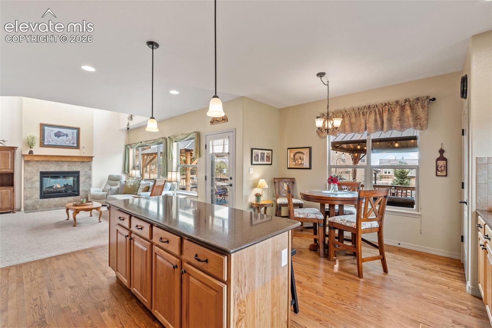 Open-concept living space featuring a central kitchen island with countertop, wood-finish flooring, a fireplace with stone surround, a dining area with a window, and recessed lighting