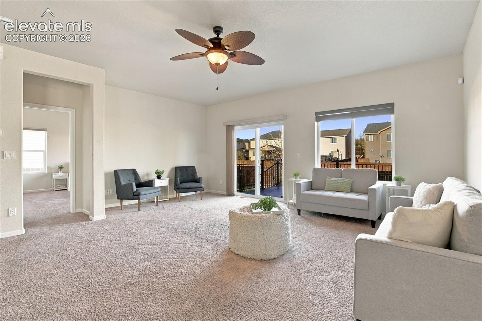 Neutral-toned carpeted flooring, central ceiling fan with integrated lighting, sliding glass door, and large windows providing natural light