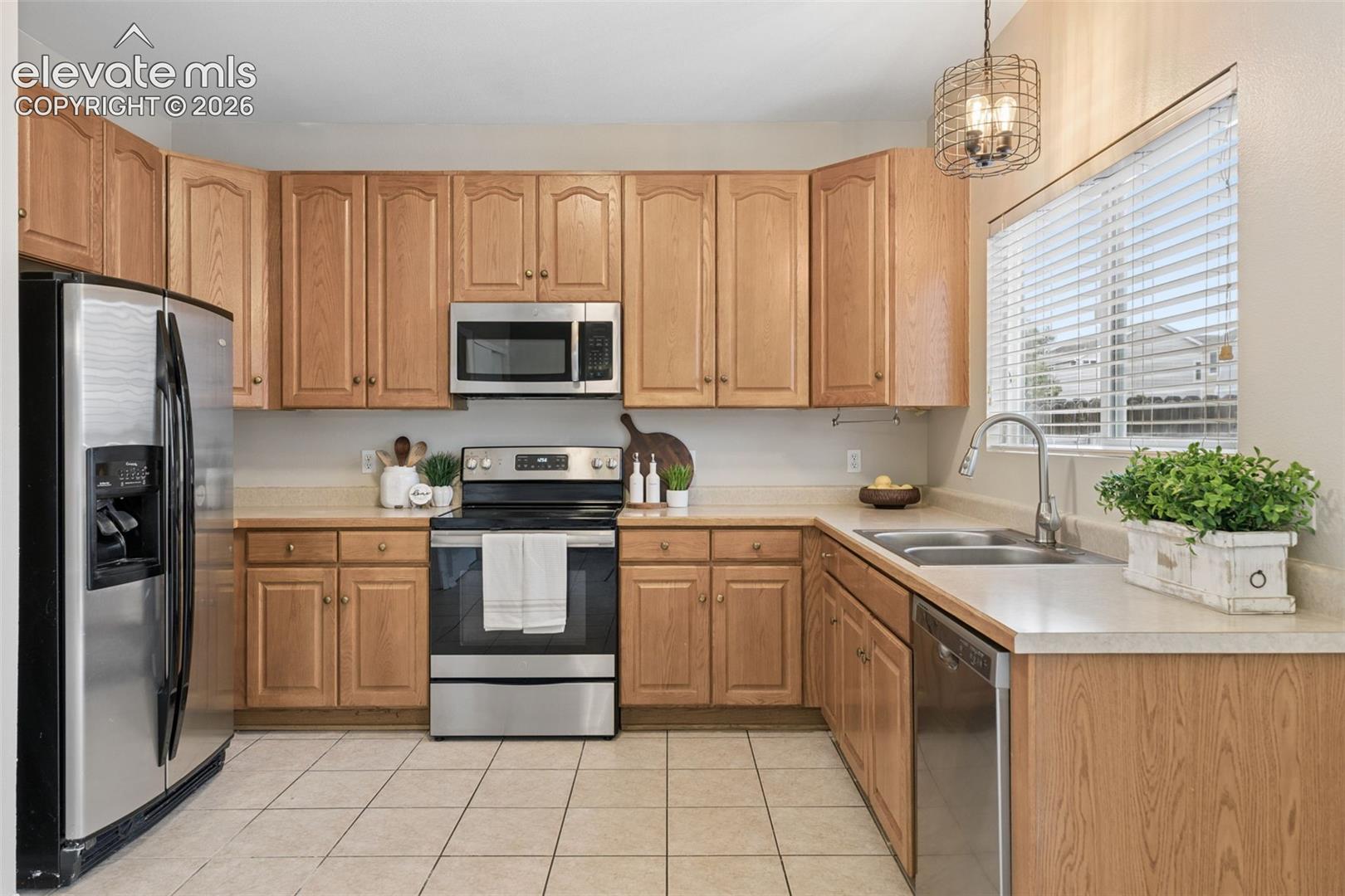 Kitchen featuring light wood cabinetry, stainless steel appliances, neutral solid-surface countertops, tile flooring, and a wire-frame pendant light fixture