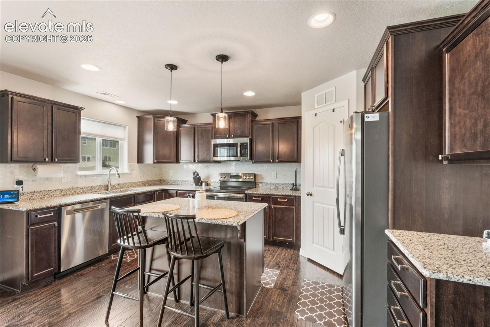Kitchen featuring dark wood-finish cabinetry, granite countertops, stainless steel appliances, a center island with pendant lighting, and wood-finish flooring
