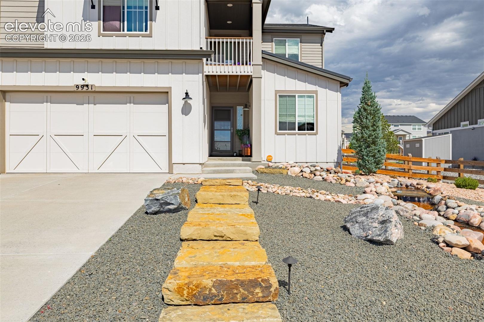Two-story exterior featuring vertical board-and-batten siding, a covered entryway with a second-story balcony, a two-car garage with carriage-style doors, and xeriscaped front yard with stone-stepped pathway and dry creek bed