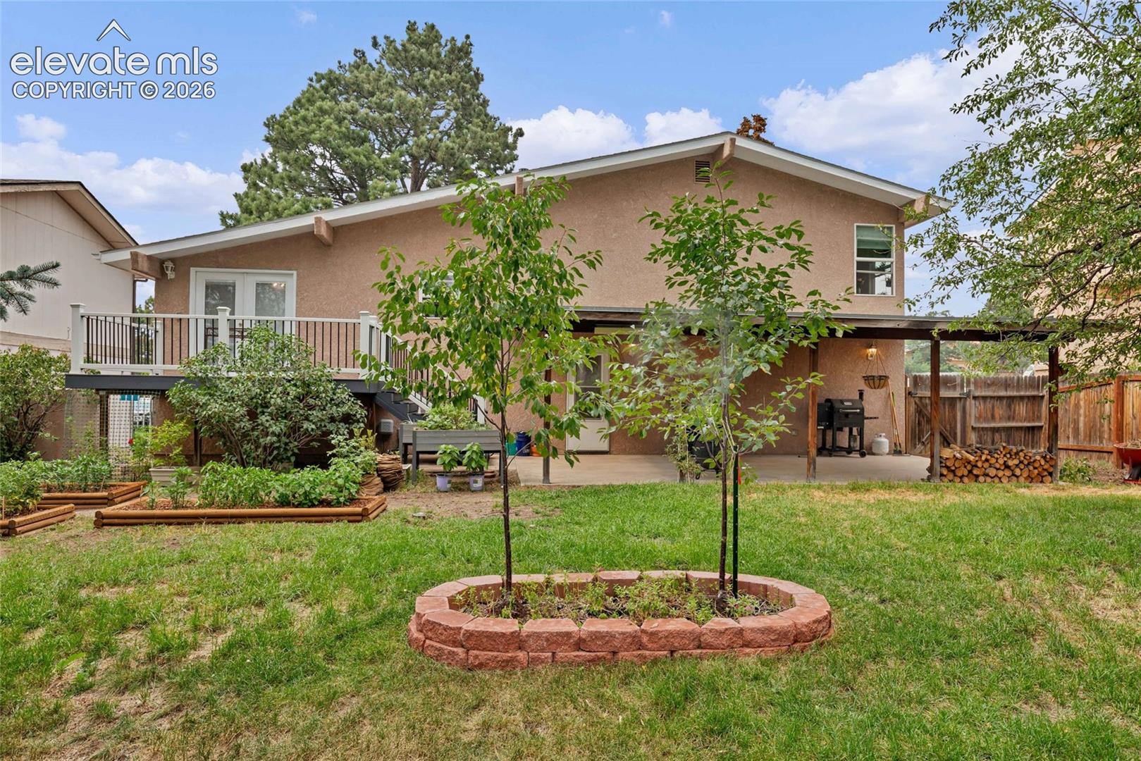 Expansive backyard featuring a stucco exterior, raised deck with white railing, covered patio, and multiple garden beds
