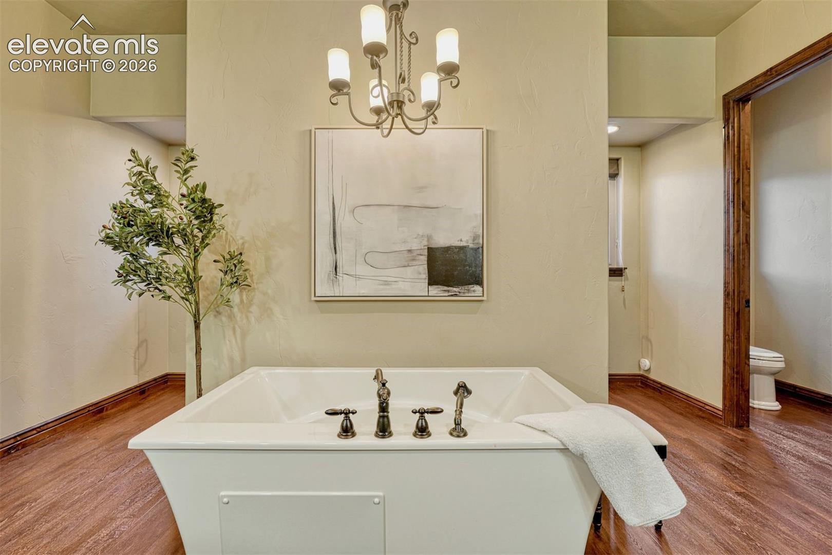 Freestanding soaking tub with bronze-finish fixtures, complemented by wood-finish flooring and a decorative chandelier