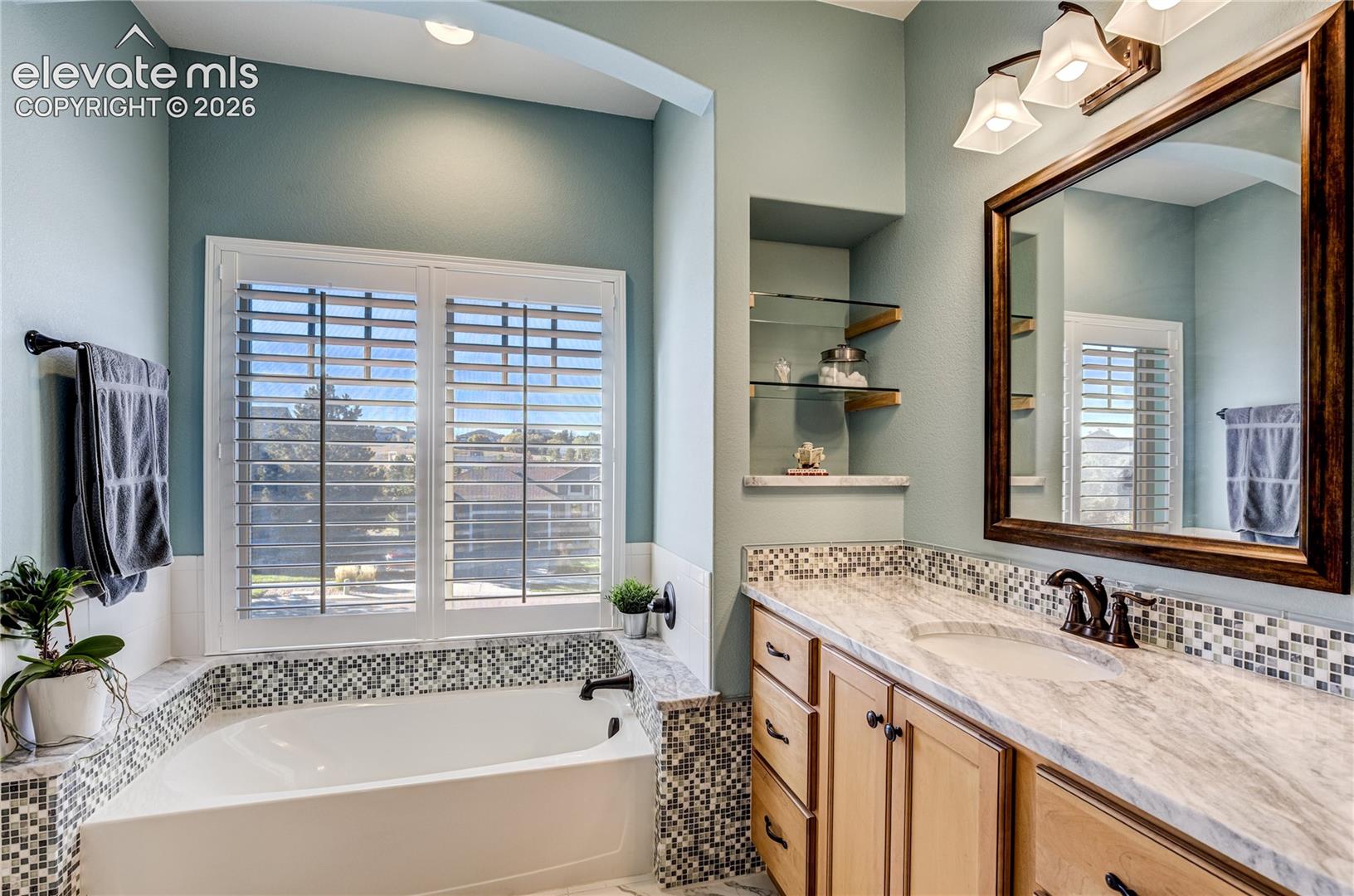 The beautifully appointed upper-level five-piece bath features a separate soaking tub set beneath plantation shutters, offering a relaxing retreat complemented by elegant tilework + thoughtful detailing.