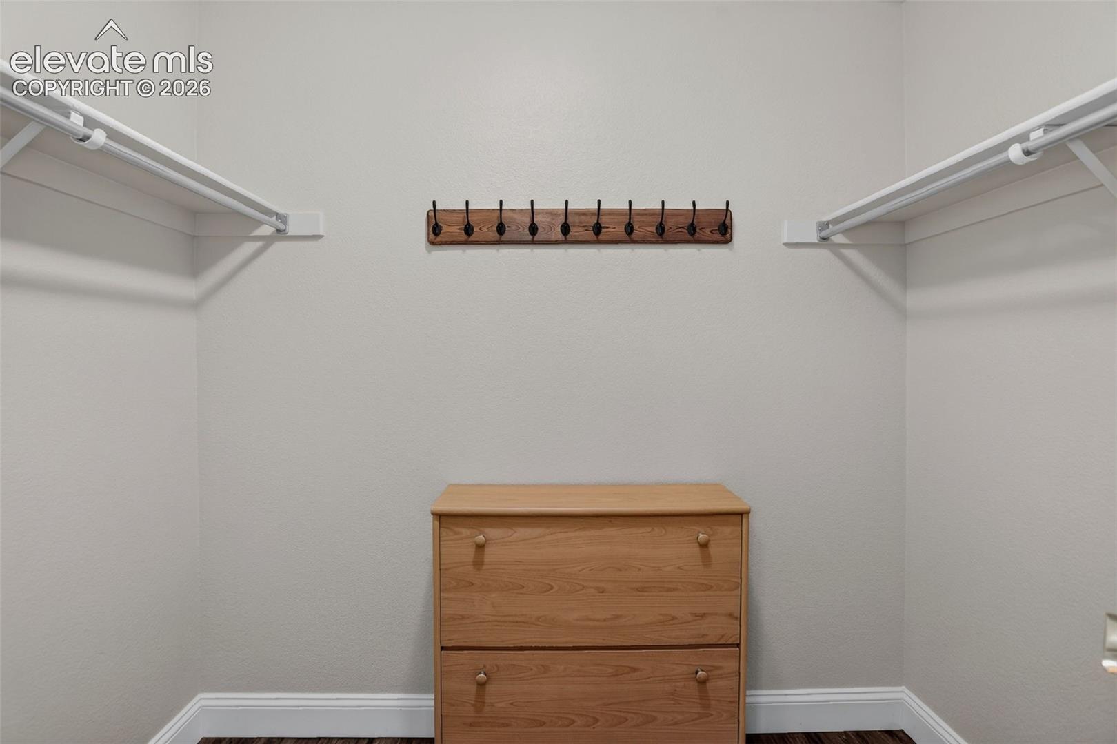 Walk-in closet with white walls, wood-finish flooring, white shelving and hanging rods, and a wood-finish coat rack