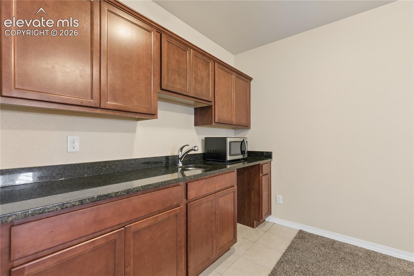 Brown wood-finish cabinetry, dark stone countertops, undermount sink, and tile flooring