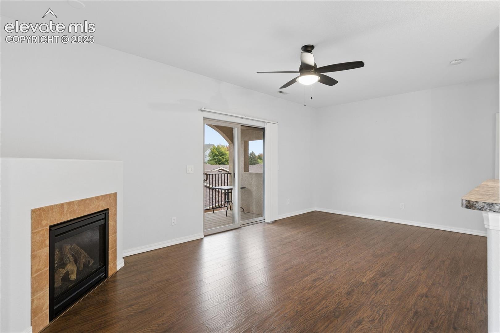 Wood-finish flooring, fireplace with neutral tile surround, ceiling fan with integrated lighting, and sliding glass door leading to a private balcony