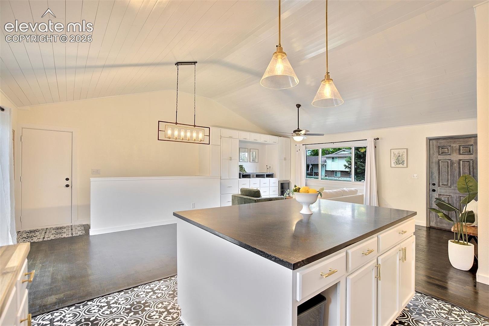 Kitchen featuring a central island with a dark countertop, white cabinetry, vaulted ceilings with beadboard detailing, decorative patterned tile flooring, and multi-light pendant fixtures