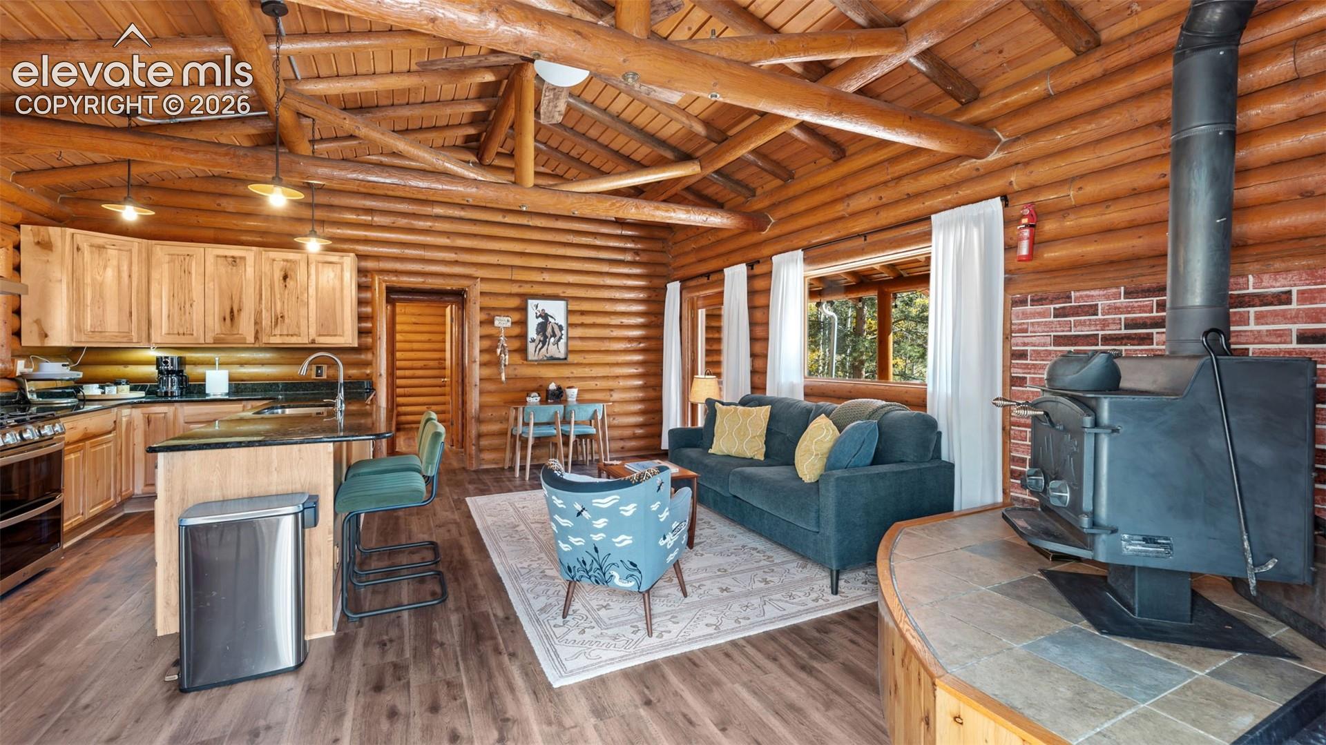 Exposed log walls, vaulted wood-beam ceiling, freestanding wood-burning stove on a tiled hearth, and integrated kitchen cabinetry