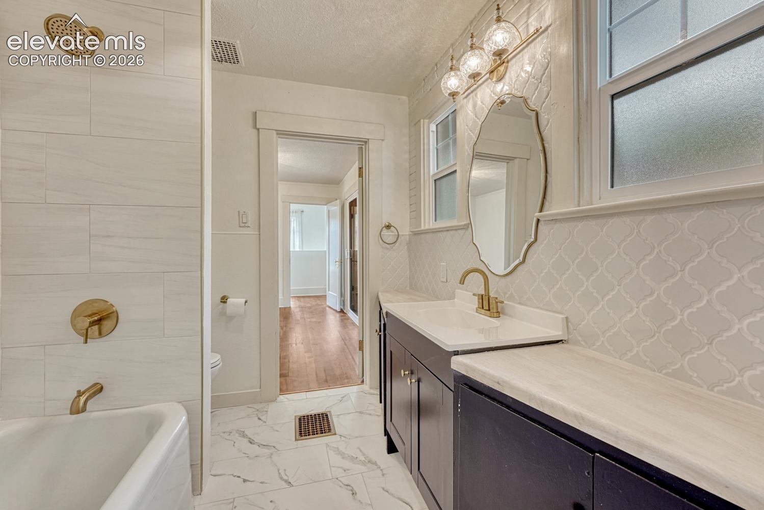 White subway tile shower surround with brass fixtures, a freestanding bathtub, marble-look floor tiling, dark wood vanity with a white countertop and brass faucet, and an arabesque tile backsplash