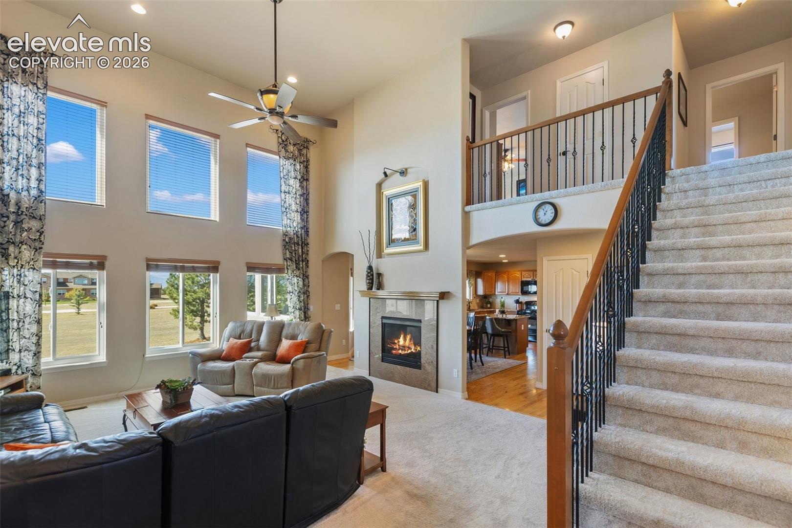 High-ceiling living area featuring a fireplace with stone-style surround, double-height windows, a ceiling fan with integrated lighting, and a staircase with wrought iron balusters