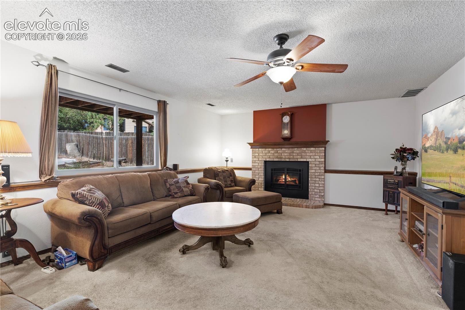 Carpeted living area featuring a brick fireplace with a wood mantle, ceiling fan with integrated lighting, and a large sliding glass door