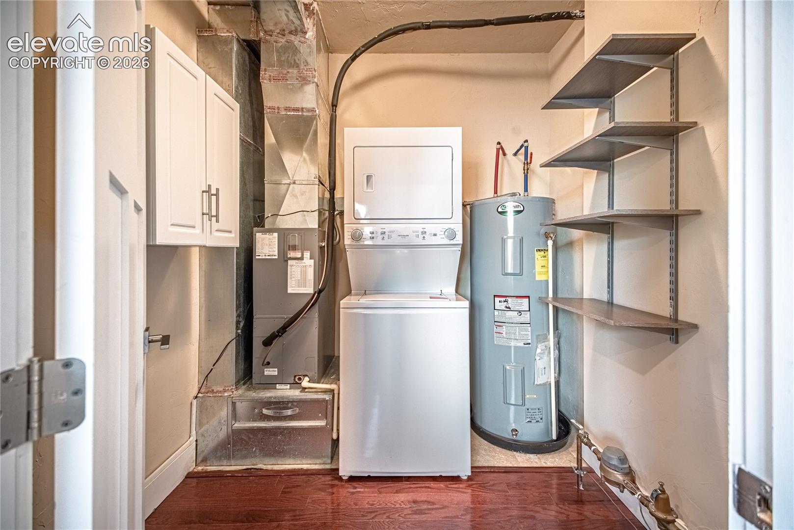 Dedicated utility area featuring wood-finish flooring, a stacked laundry appliance, a water heater, built-in wall shelving, and overhead cabinetry