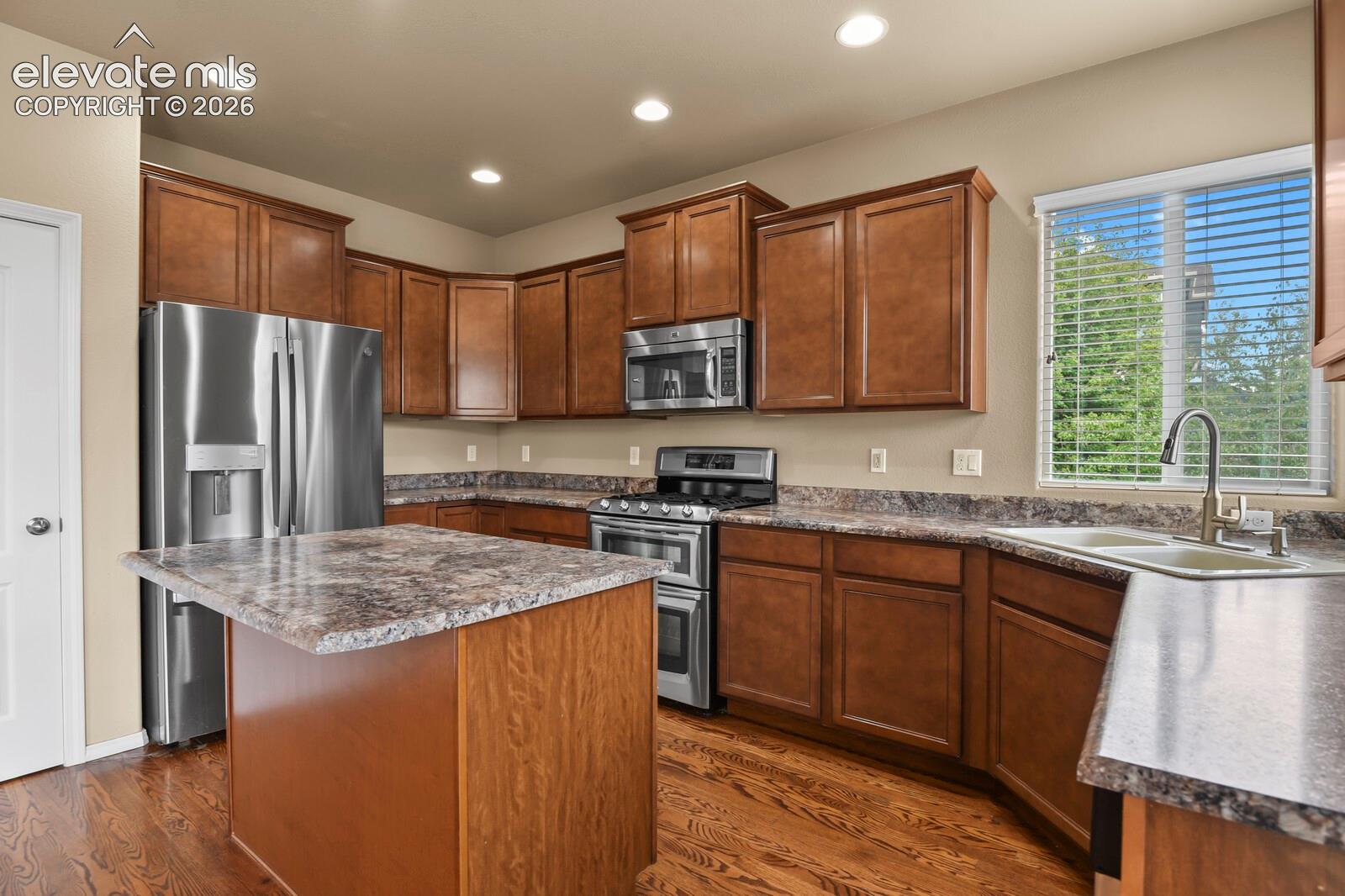 Kitchen featuring rich wood cabinetry, stainless steel appliances, a center island with countertop seating, a dual basin sink with a gooseneck faucet, and wood-finish flooring