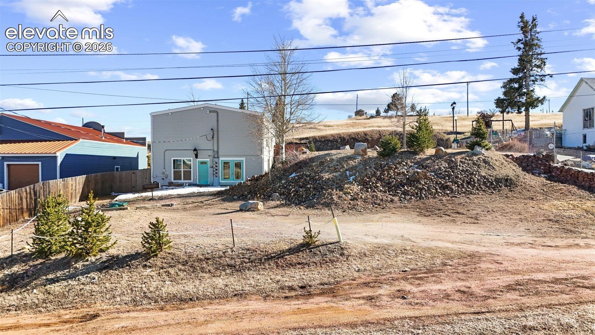 Exterior featuring gray siding, a bright blue entry door, large windows, and a raised earth mound with rock retaining elements and pine trees