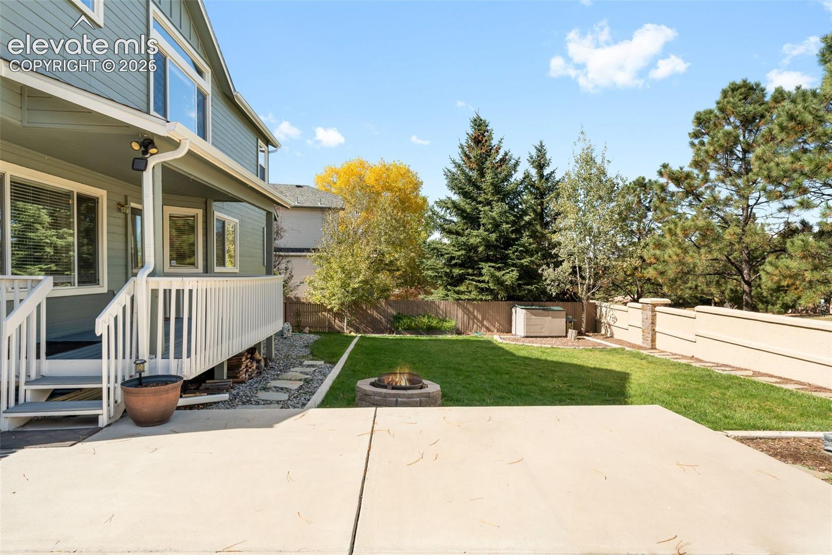 Fenced backyard featuring a raised wooden deck, concrete patio, stone fire pit, and mature perimeter trees
