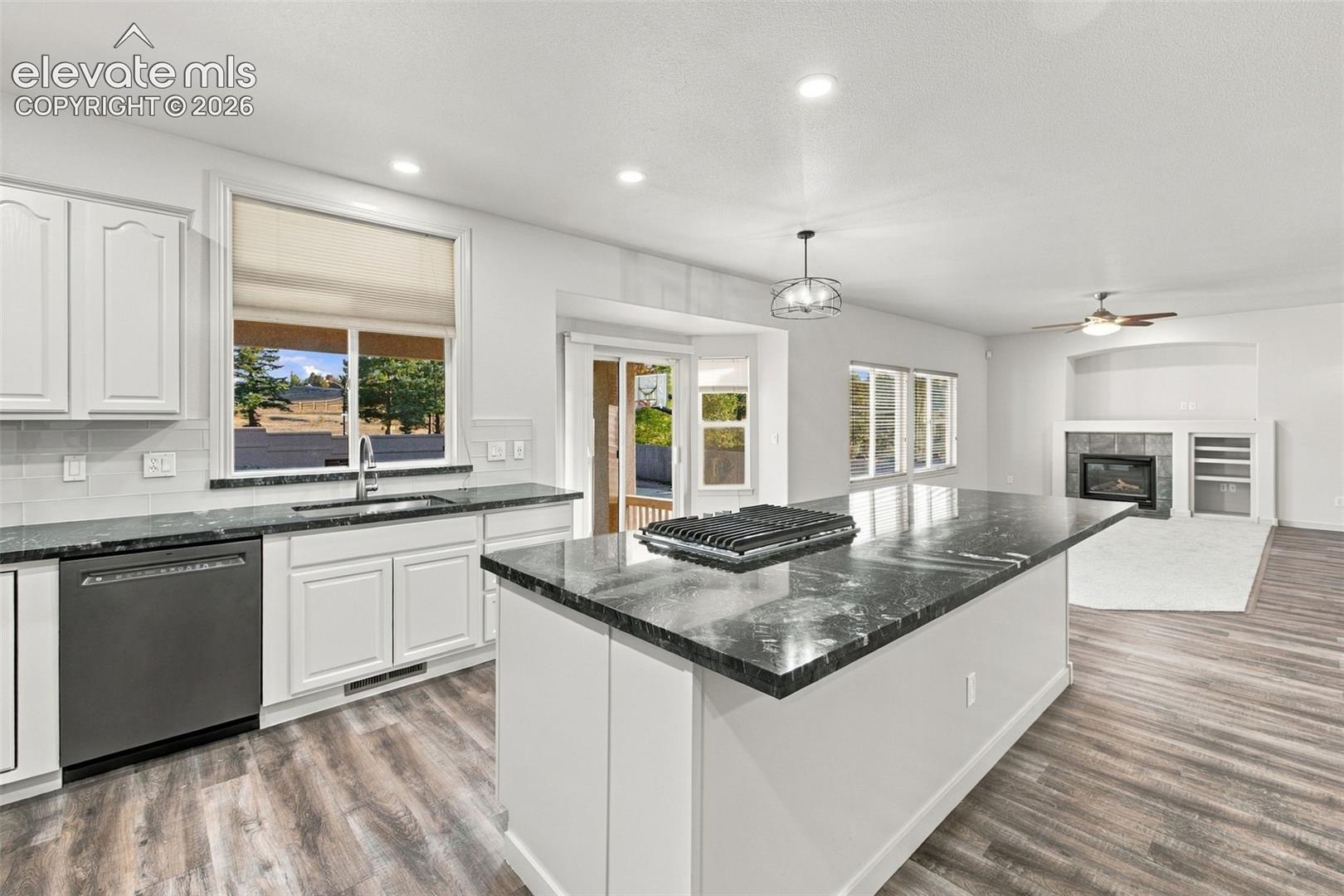 Kitchen featuring white cabinetry, dark stone countertops, an expansive center island with a built-in cooktop, wood-finish flooring, and a stone-surround fireplace