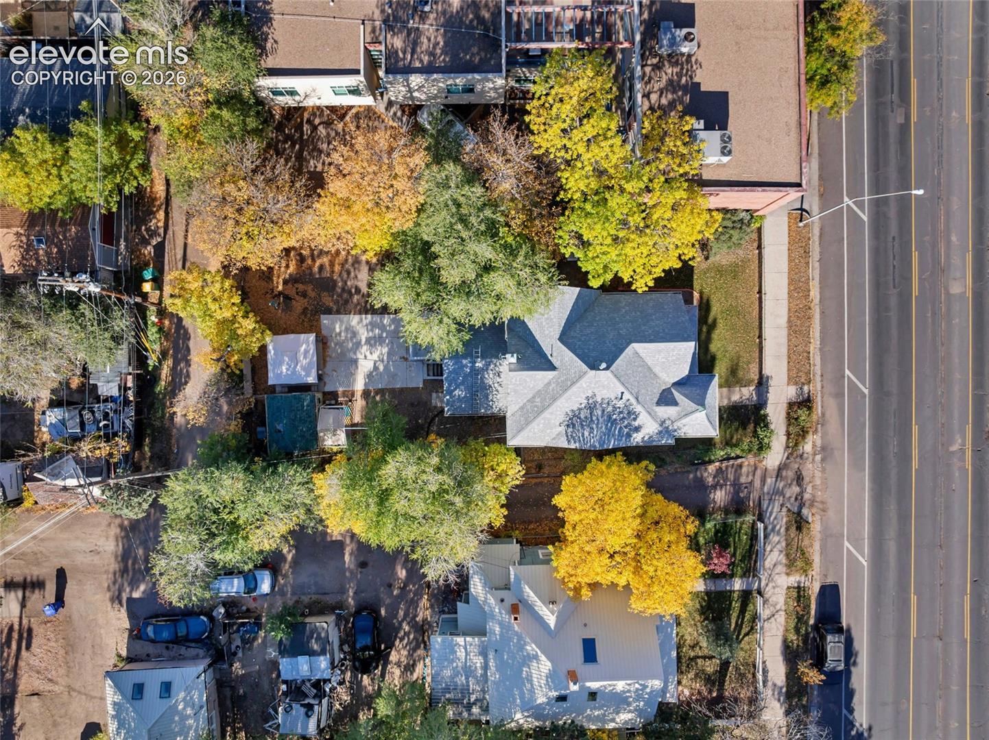 Aerial view featuring a multi-pitched shingled roof, paved patio area, and mature trees across a landscaped lot