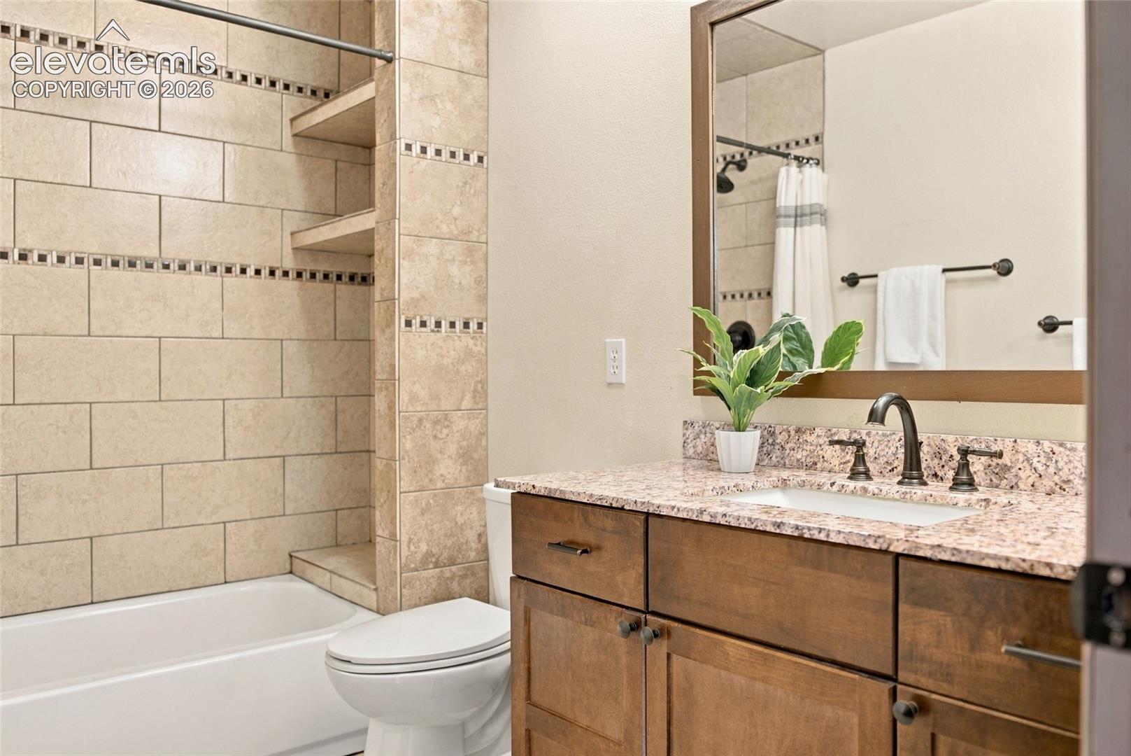 Tub-shower combination with tiled surround, built-in shelving, wood-finish vanity with stone countertop, and an integrated sink