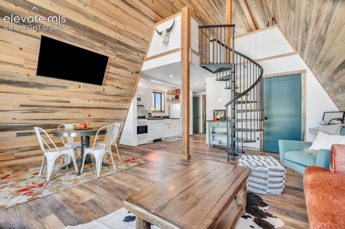 A-frame interior featuring natural wood plank wall paneling, wood-finish flooring, a metal spiral staircase, and kitchen cabinetry with a farmhouse-style sink