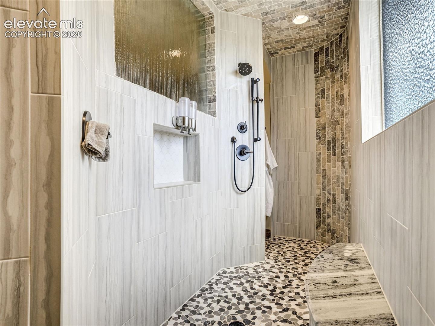 A richly detailed walk-in shower elevates the primary bath experience with pebble stone flooring, artisan tilework, bench seating, and dual shower fixtures including an overhead rain shower.