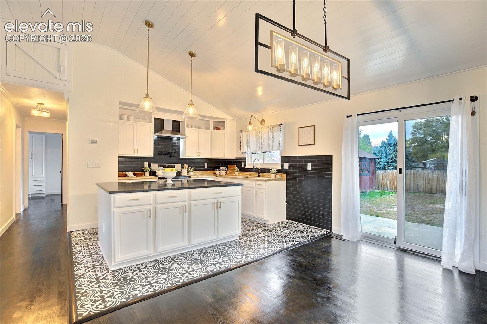 Kitchen featuring a central island with a dark countertop, white cabinetry, patterned tile flooring, contrasting black subway tile backsplash, and a vaulted ceiling with integrated pendant and rectangular chandelier lighting