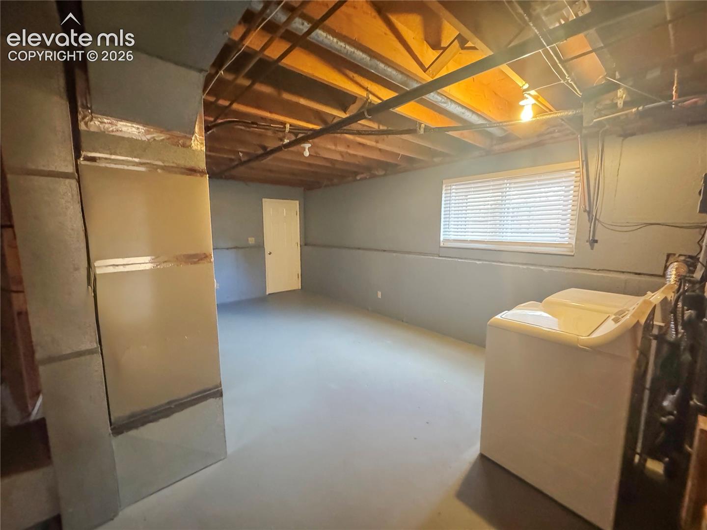 Lower level featuring painted concrete flooring, exposed wood joists, a horizontal window with blinds, and utility access