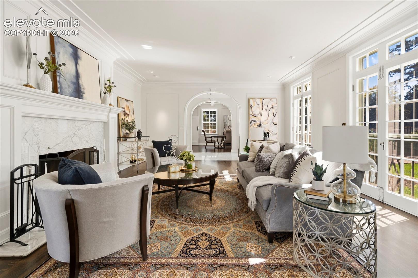 Living room featuring crown molding, wainscoting, fireplace with stone surround, arched architectural detailing, and French doors