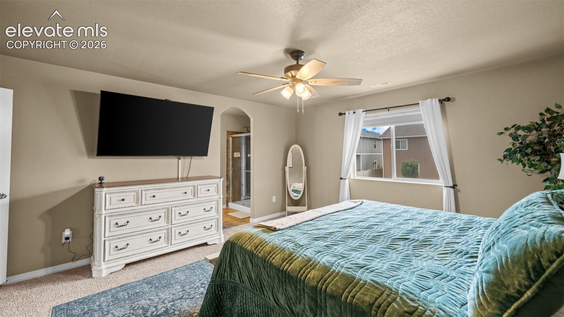 Carpeted bedroom featuring a ceiling fan with light fixture, a window with white trim, and a doorway leading to a bathroom with a glass-enclosed shower