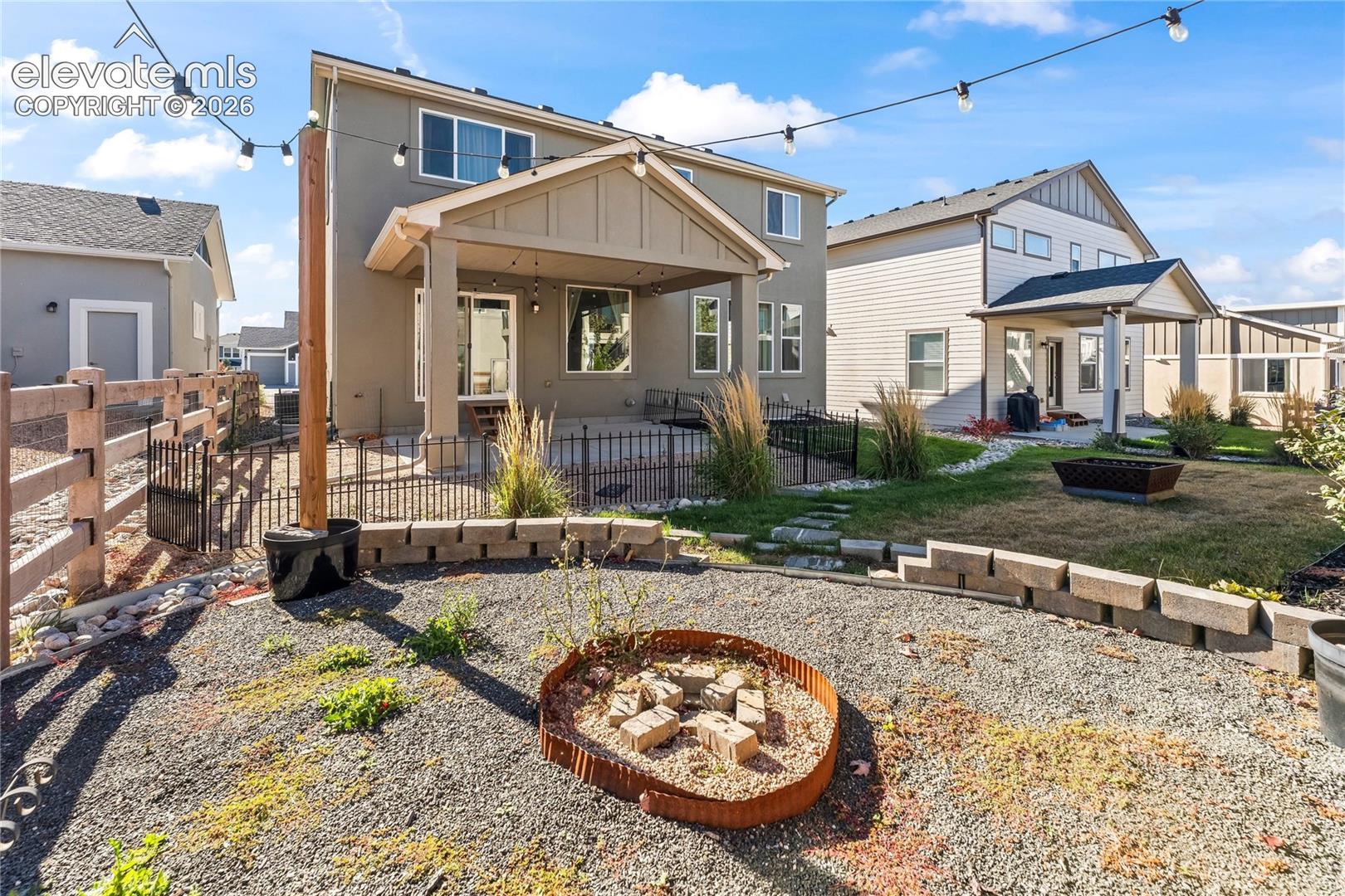 Covered patio with integrated string lighting, ornamental iron fencing, multi-level stone block retaining walls, and circular gravel fire pit area
