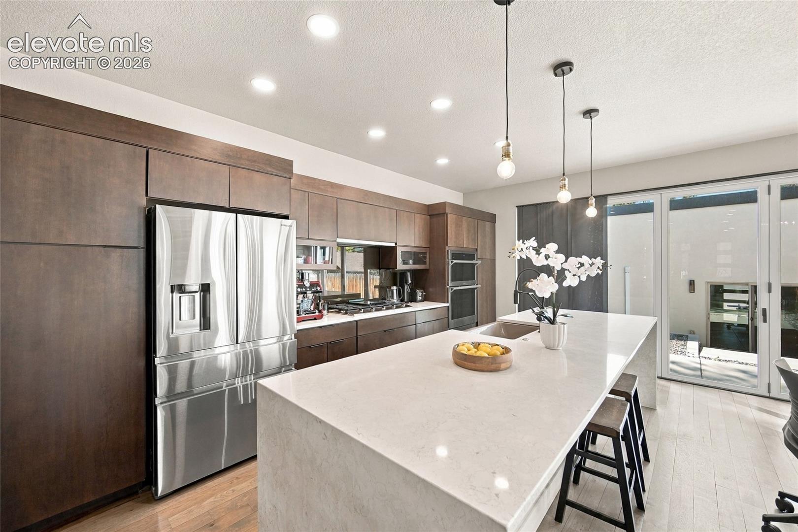 Kitchen featuring flat-panel cabinetry, a waterfall-edge island with stone countertop, stainless steel appliances, recessed ceiling lighting, and pendant fixtures