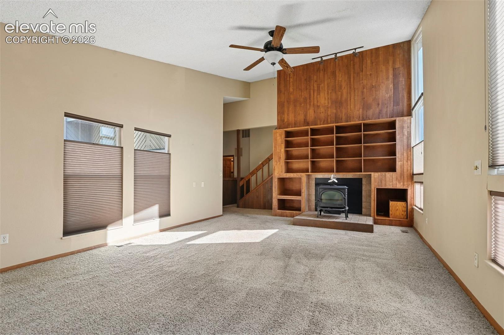Vaulted ceilings featuring a ceiling fan, floor-to-ceiling wood paneling, built-in shelving, a wood-burning stove on a raised hearth, and tall multi-pane windows