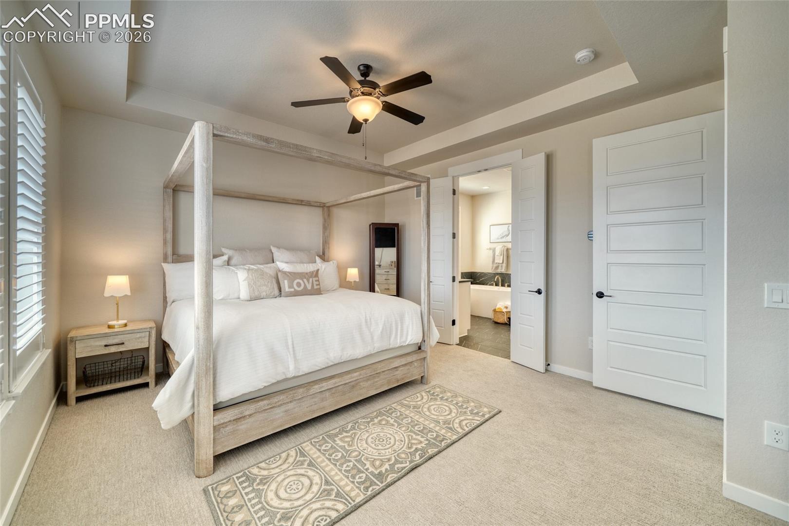 Bedroom with a tray ceiling, light colored carpet, ensuite bathroom, and ceiling fan