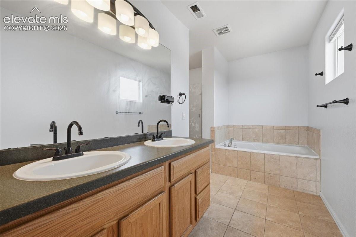 Dual vanity with undermount sinks and black fixtures, expansive countertop space, built-in bathtub with tiled surround, and tiled flooring